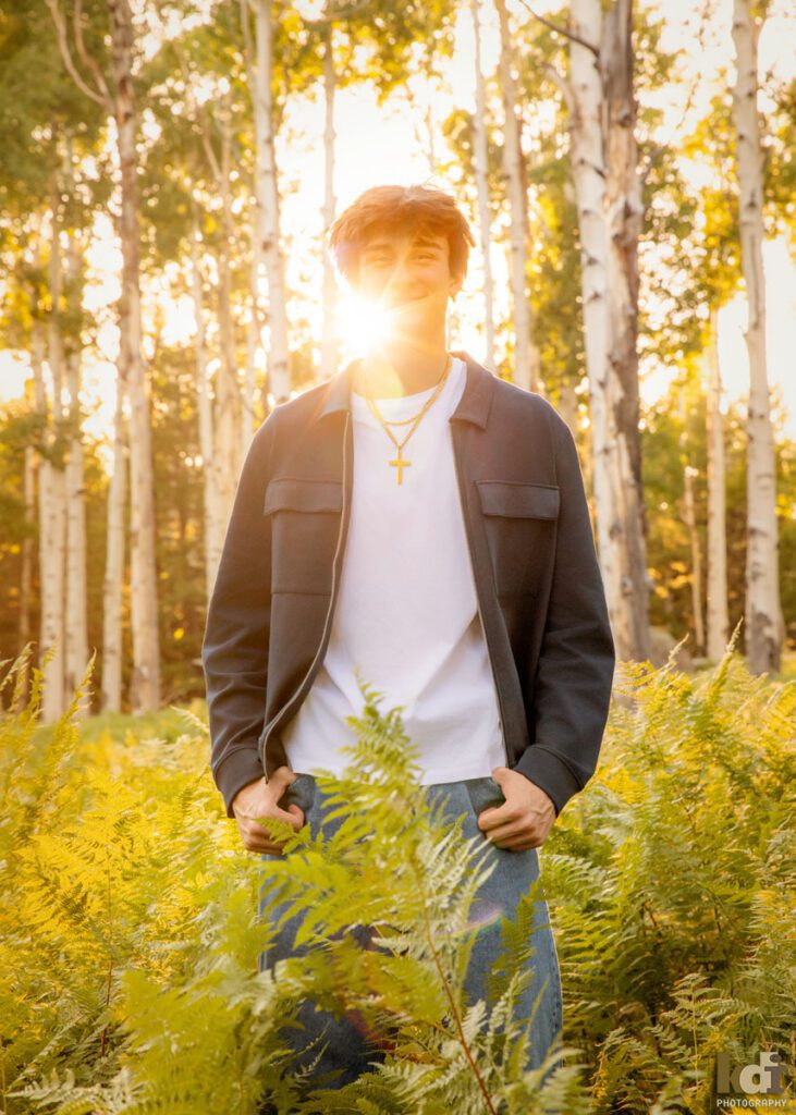 High school senior photos showing boy with brown hair, wearing white t-shirt and blue jacket, smiling at the camera, back lit with flare from the setting sun, among the summer ferns and aspen trees, portrait photography in Flagstaff AZ by senior photographer KDI Photography