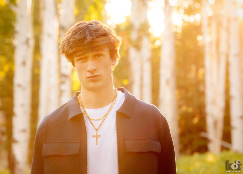 High school senior photos showing boy with brown hair, wearing white t-shirt and blue jacket, making a funny face at the camera, back lit but the setting sun, among the summer ferns and aspen trees, portrait photography in Flagstaff AZ by senior photographer KDI Photography