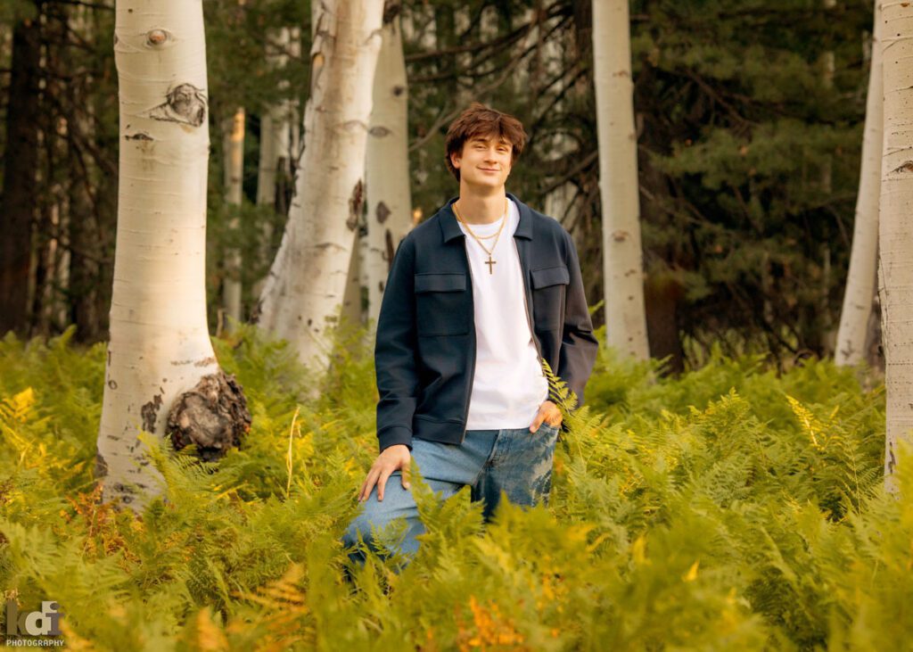 High school senior photos showing boy with brown hair, wearing white t-shirt and blue jacket,  among the summer ferns and aspen trees, portrait photography in Flagstaff AZ by senior photographer KDI Photography