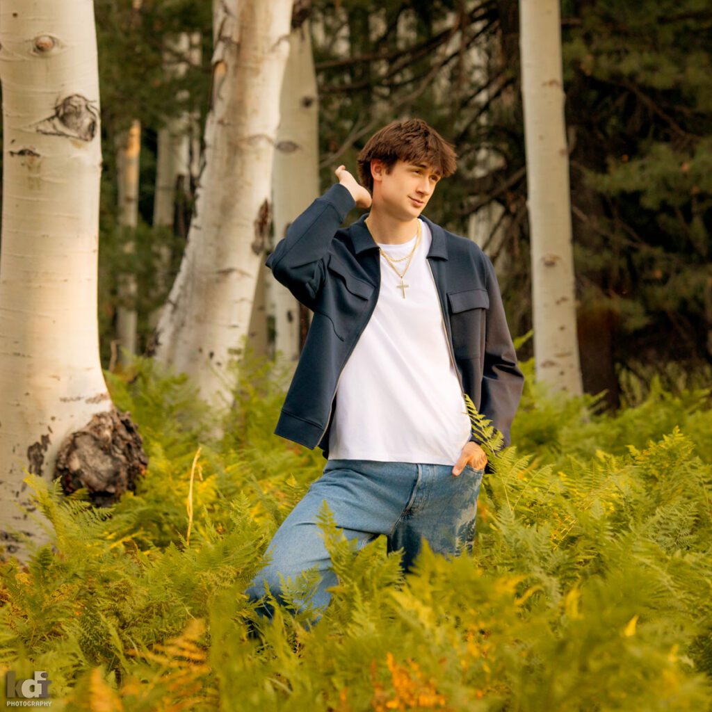 High school senior photos showing boy with brown hair, wearing white t-shirt and blue jacket, making a funny gesture with his hair among the summer ferns and aspen trees, portrait photography in Flagstaff AZ by senior photographer KDI Photography