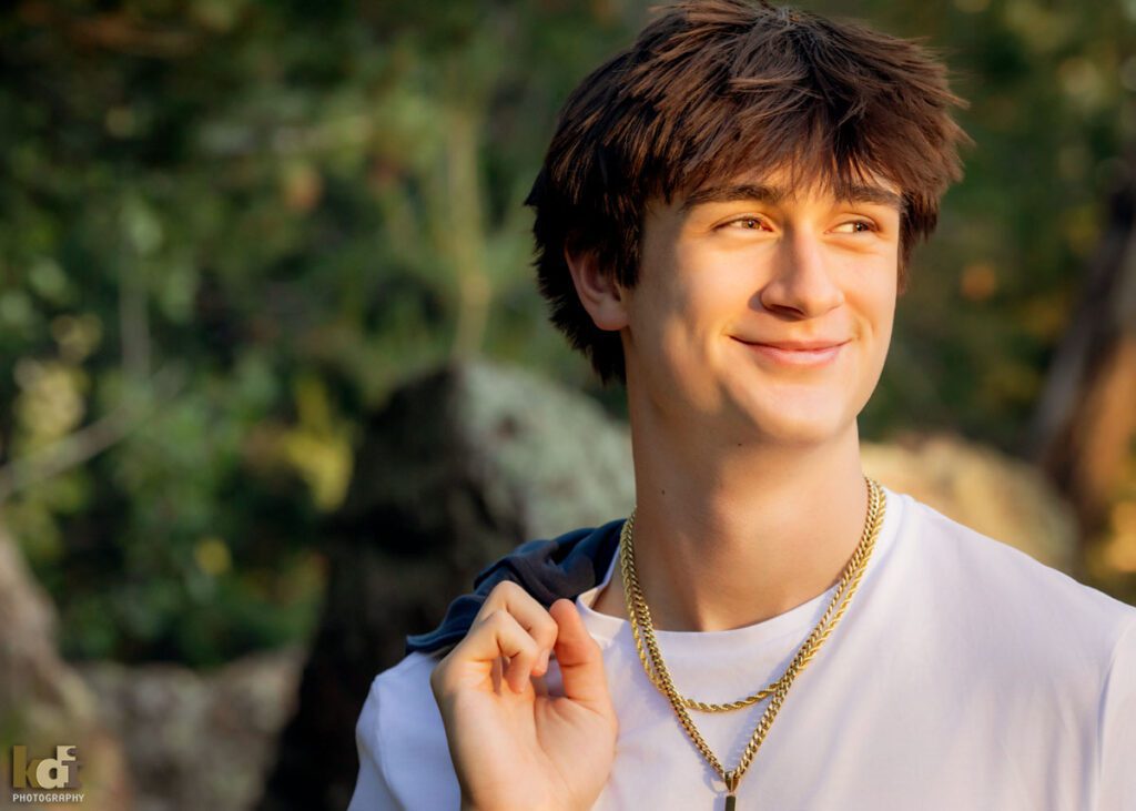 High school senior photos showing boy with brown hair, wearing white t-shirt and holding a blue jacket, smiling off camera, among the summer ferns and aspen trees, portrait photography in Flagstaff AZ by senior photographer KDI Photography