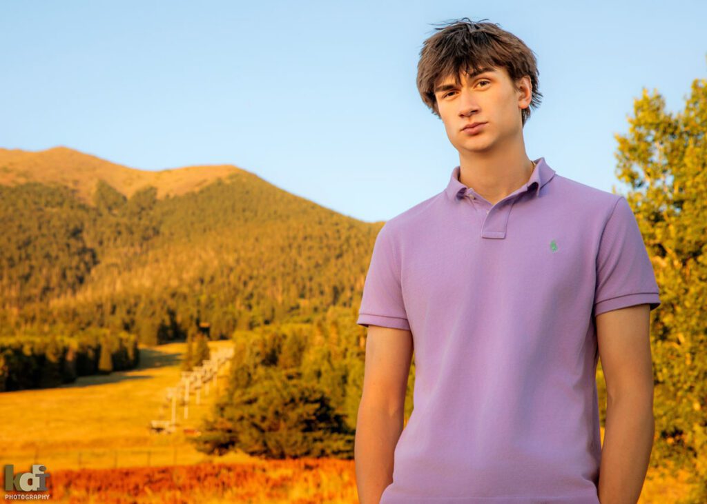 High school senior photos showing boy with brown hair, making a funny face and wearing a purple collared t-shirt, with the San Francisco Peaks in the background, portrait photography in Flagstaff AZ by senior photographer KDI Photography