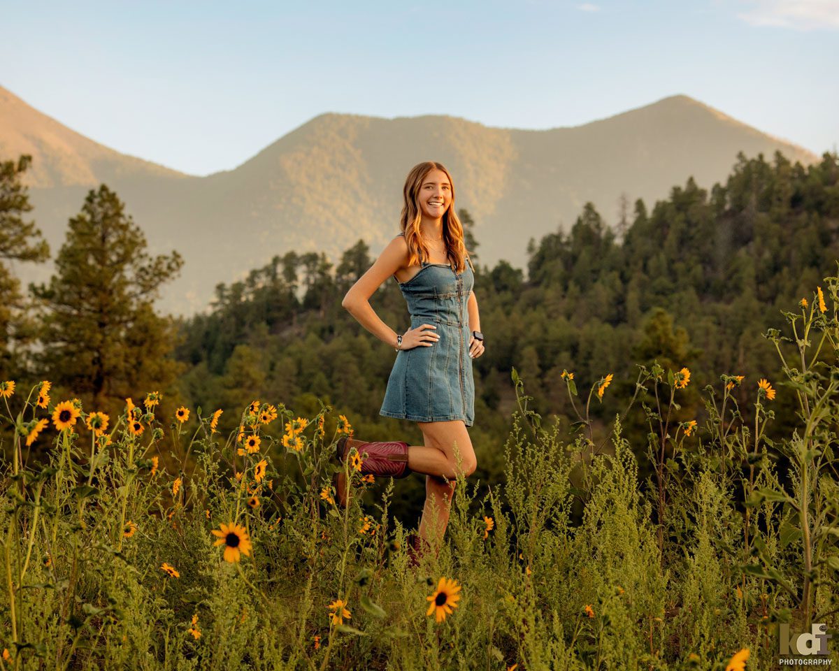 Senior photo of blonde girl in a denim dress, kicking up her cowboy boots, in the wildflowers with the San Francisco Peaks in the background, location senior portraits by Flagstaff photographer, KDI Photography. 
