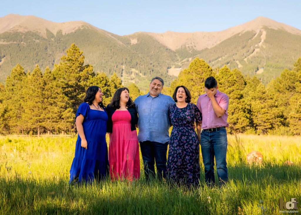 Family photos of 2 parents and 3 grown children, laughing at each other in the summer grass, with the San Francisco Peaks behind them, family portraits by Flagstaff photographer KDI Photography