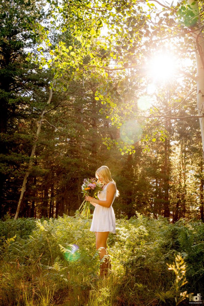 High school senior photos showing girl with blond hair, in white dress and cowboy boots, standing in the aspen trees, holding and inhaling from a bouquet of flowers, portrait photography in Flagstaff AZ by senior photographer KDI Photography
