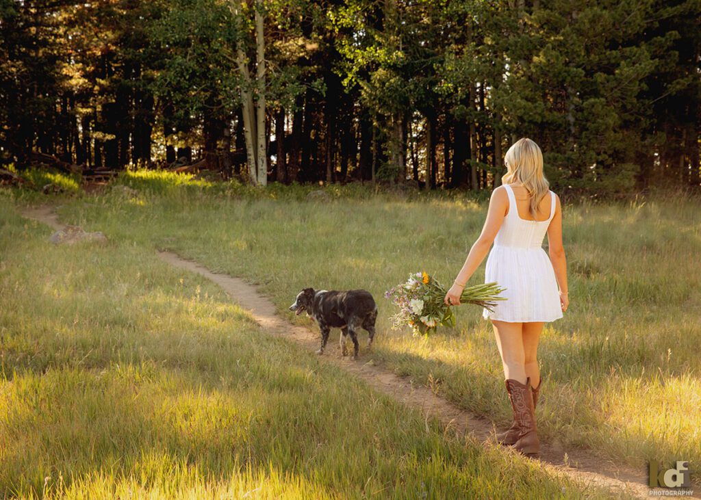 High school senior photos showing girl with blond hair, in white dress and cowboy boots, walking with her dog while holding a bouquet of flowers, portrait photography in Flagstaff AZ by senior photographer KDI Photography