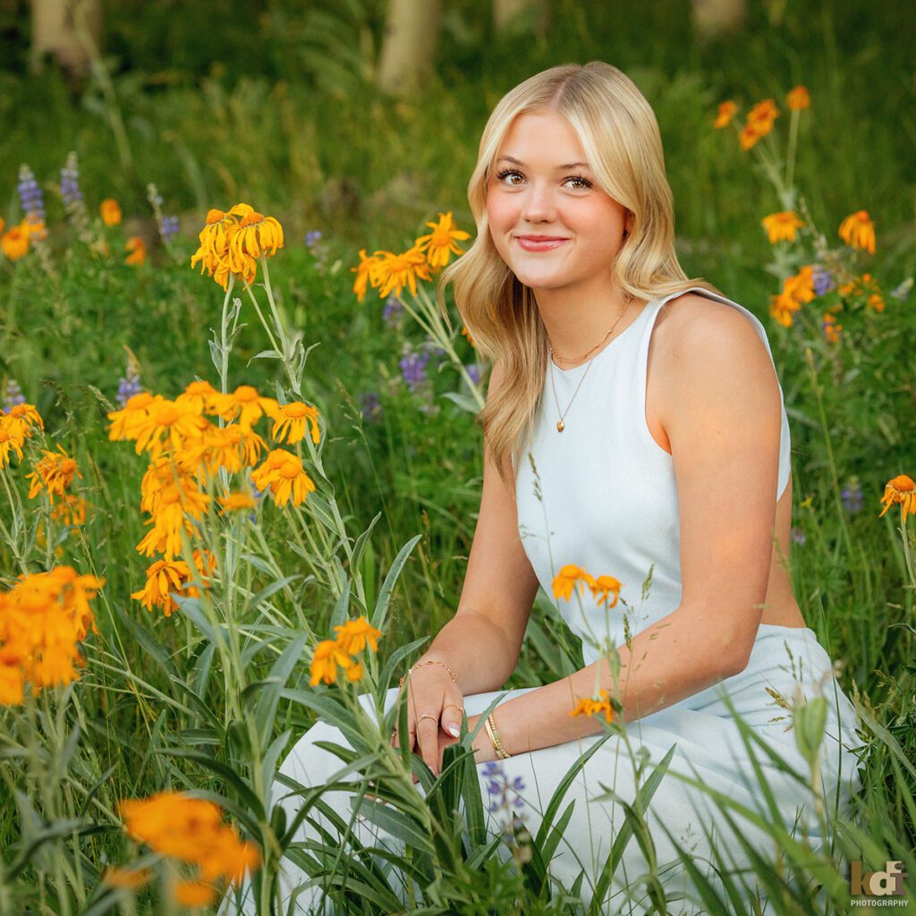 color portrait, high school senior photo showing a blonde haired girl grinning, looking at the camera, wearing in blue dress sitting in a meadow surrounded by yellow flowers, portrait photography in Flagstaff AZ by senior photographer KDI Photography