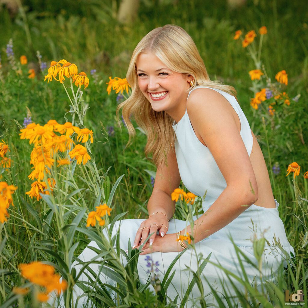 color portrait, high school senior photo showing a blonde haired girl giggling, wearing in blue dress sitting in a meadow surrounded by yellow flowers, portrait photography in Flagstaff AZ by senior photographer KDI Photography