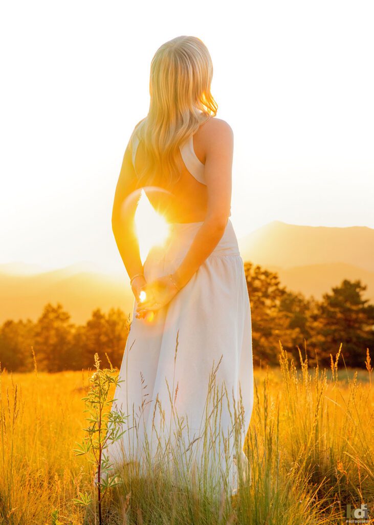 color portrait, high school senior photo showing girl with blond hair, in blue dress standing in a meadow at sunset, with sun flare shining through her arm, looking away from the camera, portrait photography in Flagstaff AZ by senior photographer KDI Photography