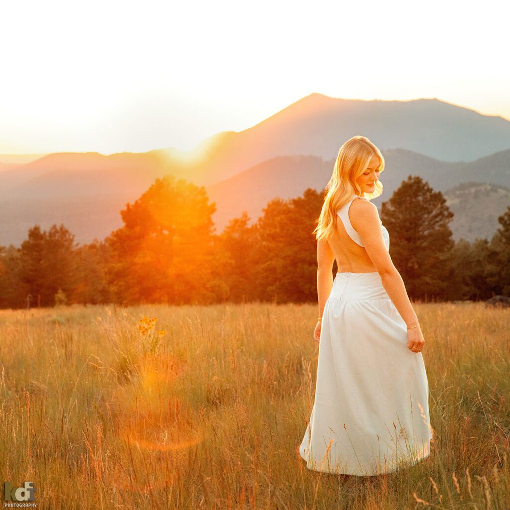 color portrait, high school senior photo showing girl with blond hair, in blue dress standing in a meadow at sunset, with sun flare from the setting sun shining through the portrait as she holds her dress, portrait photography in Flagstaff AZ by senior photographer KDI Photography