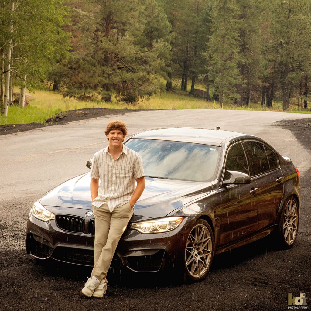 A smiling young man stands cheerfully, leaning against his car as the rain drops fall around him, senior photos on location in the San Francisco Peaks, by Flagstaff photographer, KDI Photography.