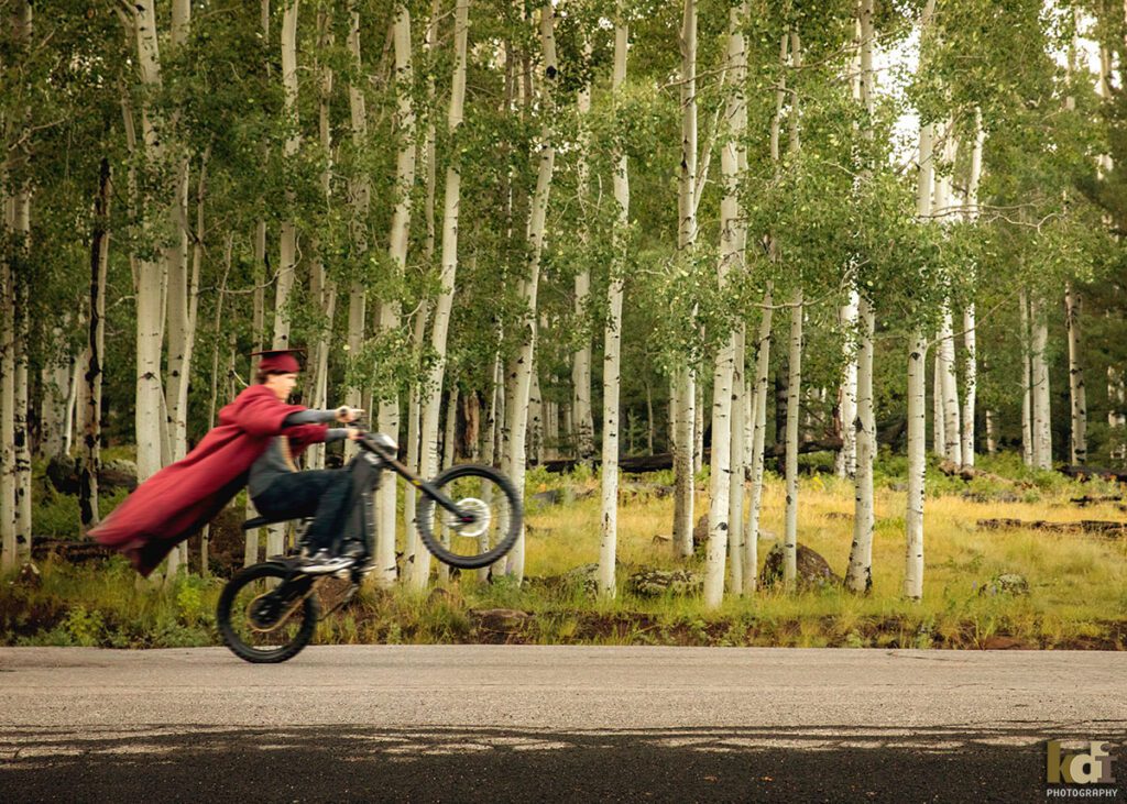 A senior boy in his cap and gown does a wheelie on his dirt bike on a road through the aspens, senior photos on location in the San Francisco Peaks, by Flagstaff photographer, KDI Photography.