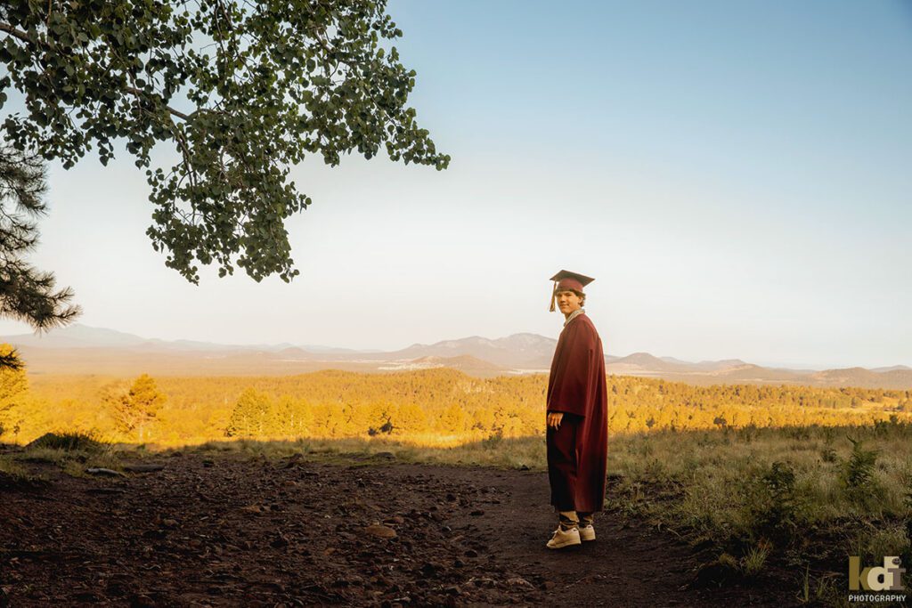 A senior boy in his cap and gown turns back to look at the camera, senior photos on location in the San Francisco Peaks, by Flagstaff photographer, KDI Photography.