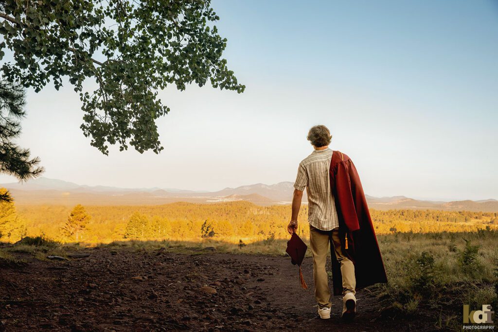 A senior boy with his graduation gown over his shoulder, holing his high school graduation cap in his hand, walks away, senior photos on location in the San Francisco Peaks, by Flagstaff photographer, KDI Photography.
