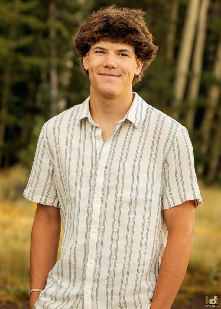 A teenage boy in a white striped short sleeved shirt, smiles at the camera while standing in the aspens, senior photos on location in the San Francisco Peaks, by Flagstaff photographer, KDI Photography.