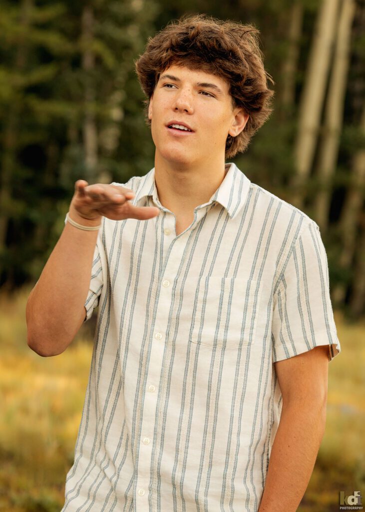 A teenage boy in a white striped short sleeved shirt, tells a story while standing in the aspens, senior photos on location in the San Francisco Peaks, by Flagstaff photographer, KDI Photography.