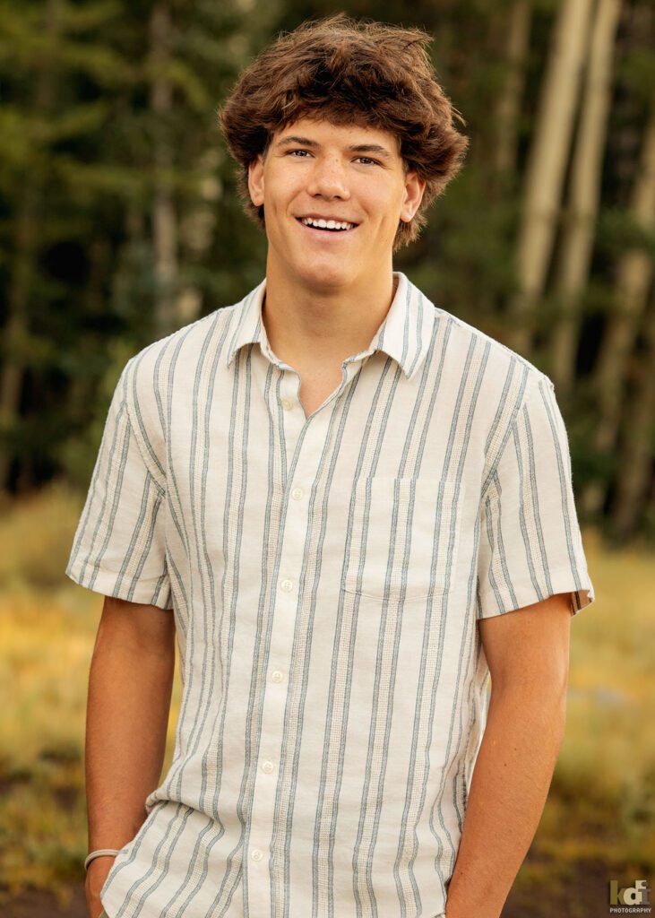 A teenage boy in a white striped short sleeved shirt, laughs at the camera while standing in the aspens, senior photos on location in the San Francisco Peaks, by Flagstaff photographer, KDI Photography.