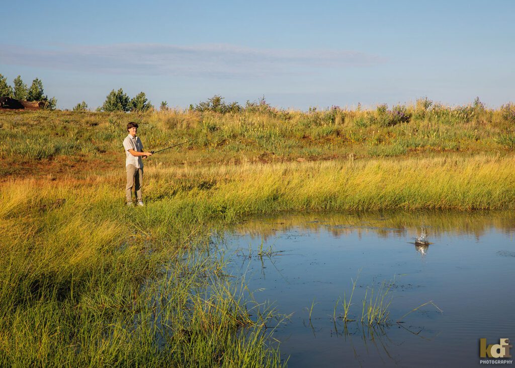 A teenage boy in a white striped shirt, casts his fishing line into an alpine pond, senior photos on location in the San Francisco Peaks, by Flagstaff photographer, KDI Photography.