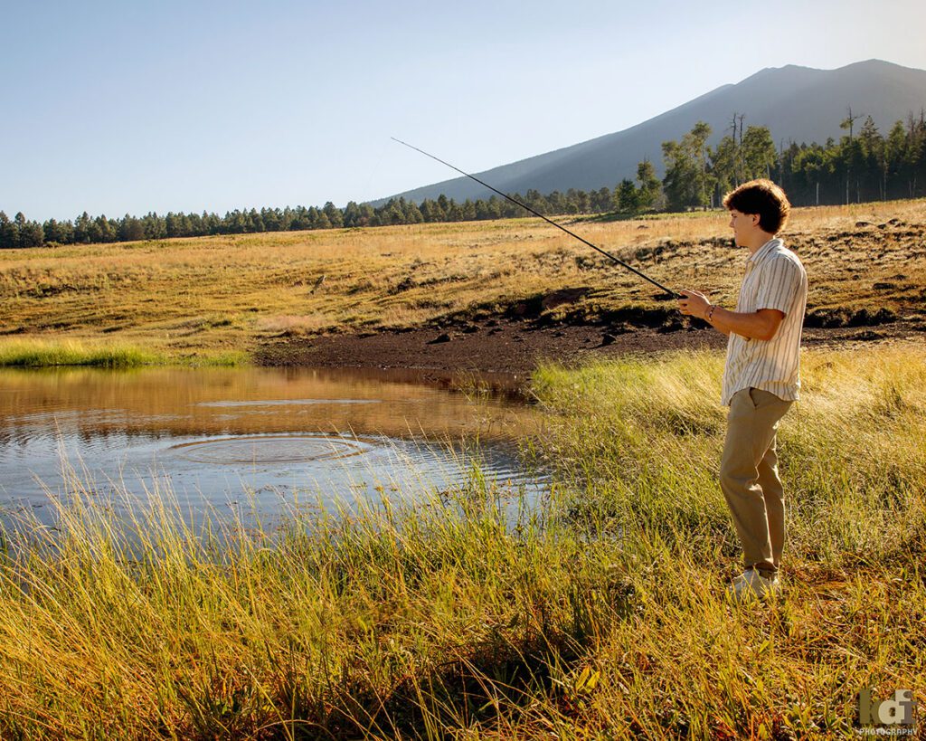 A teenage boy in a white striped shirt, casts his fishing line into an alpine pond with the mountains in the background, senior photos on location in the San Francisco Peaks, by Flagstaff photographer, KDI Photography.