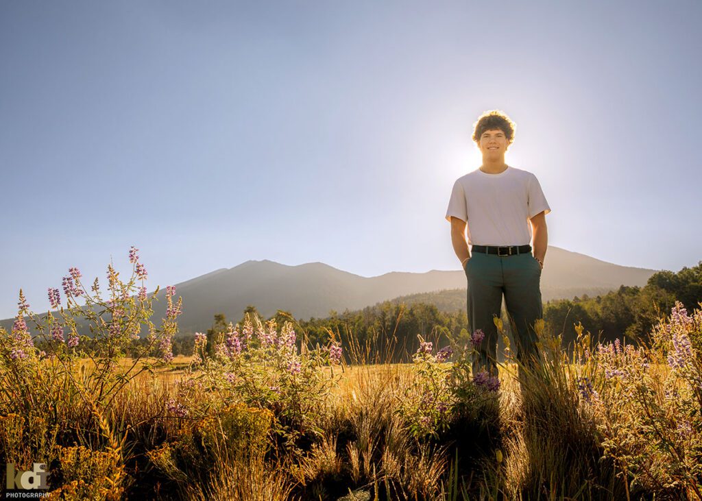 A teenage boy in a white short sleeved shirt, standing in the summer lupine and wildflowers, senior photos on location with the San Francisco Peaks in the background, by Flagstaff photographer, KDI Photography.