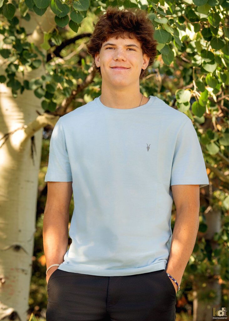A teenage boy in a blue short sleeved shirt, smiles, laughs and tells a story while standing in the aspens, senior photos on location in the San Francisco Peaks, by Flagstaff photographer, KDI Photography.