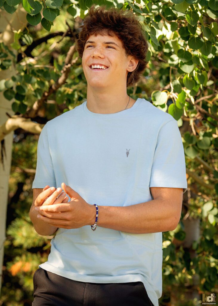 A teenage boy in a blue short sleeved shirt, smiles, laughs and tells a story while standing in the aspens, senior photos on location in the San Francisco Peaks, by Flagstaff photographer, KDI Photography.