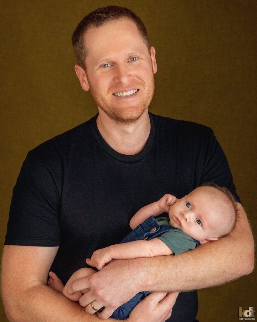 A father smiles, holding newborn baby boy, color newborn portrait of a new family in a photo studio in Flagstaff by newborn and family photographer, KDI Photography