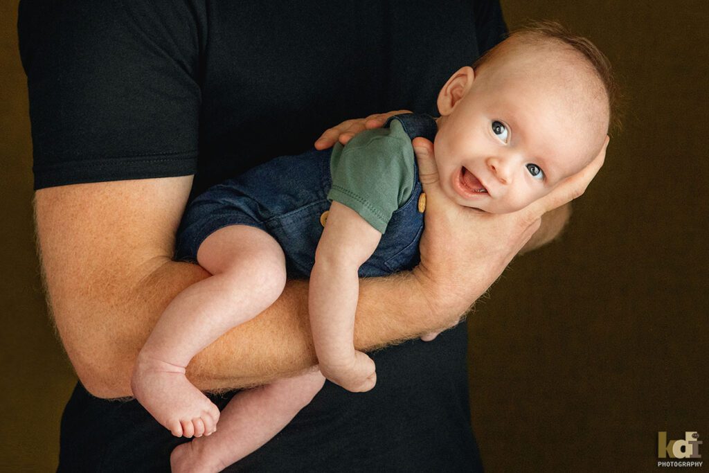 A newborn baby boy smiles as he's held in his father's arms, color newborn portrait of a new baby in a photo studio in Flagstaff by newborn and family photographer, KDI Photography