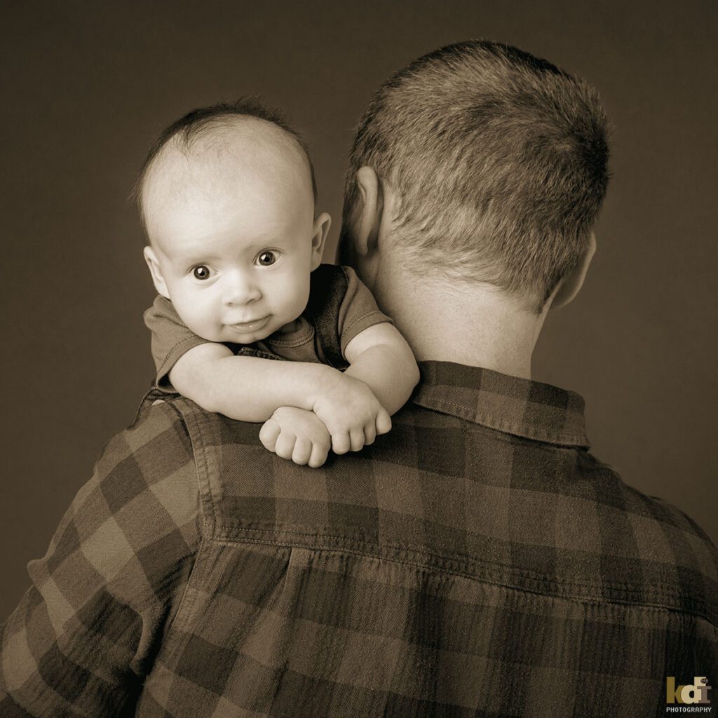 A newborn baby boy peers over his father's shoulder, black and white newborn portrait of a new family in a photo studio in Flagstaff by newborn and family photographer, KDI Photography