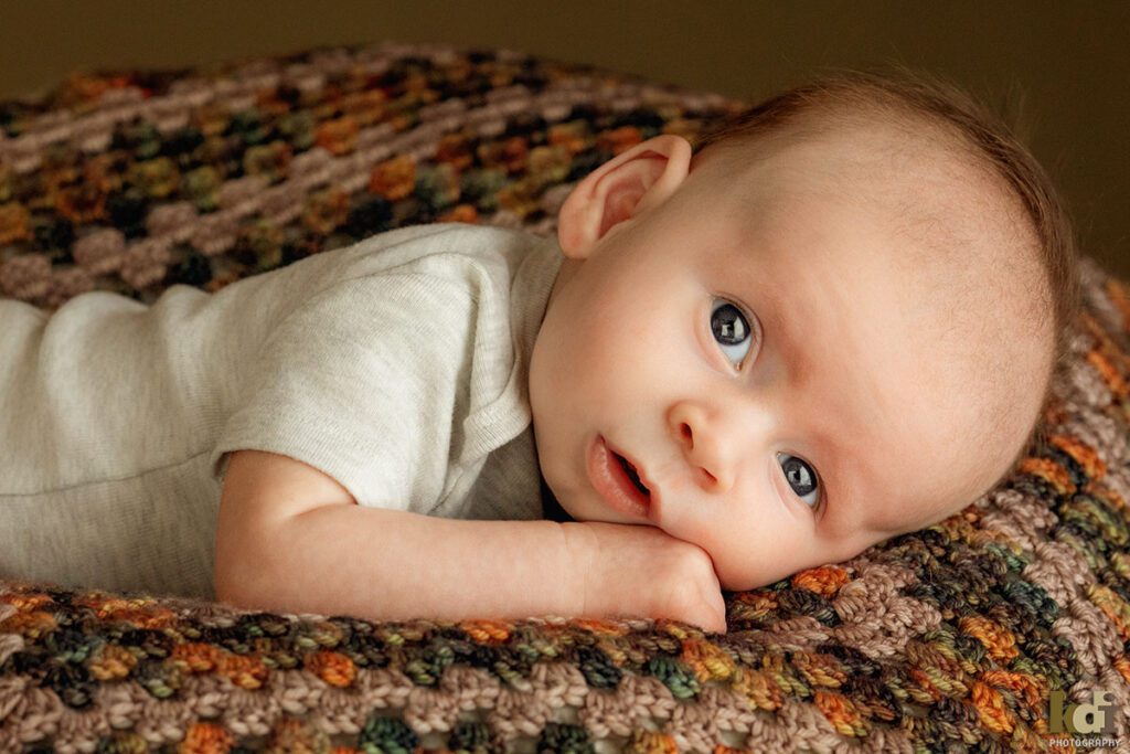 A color studio newborn portrait of a newborn baby boy looking sweetly at the camera while laying on a handmade baby blanket in a photo studio in Flagstaff by newborn and family photographer, KDI Photography