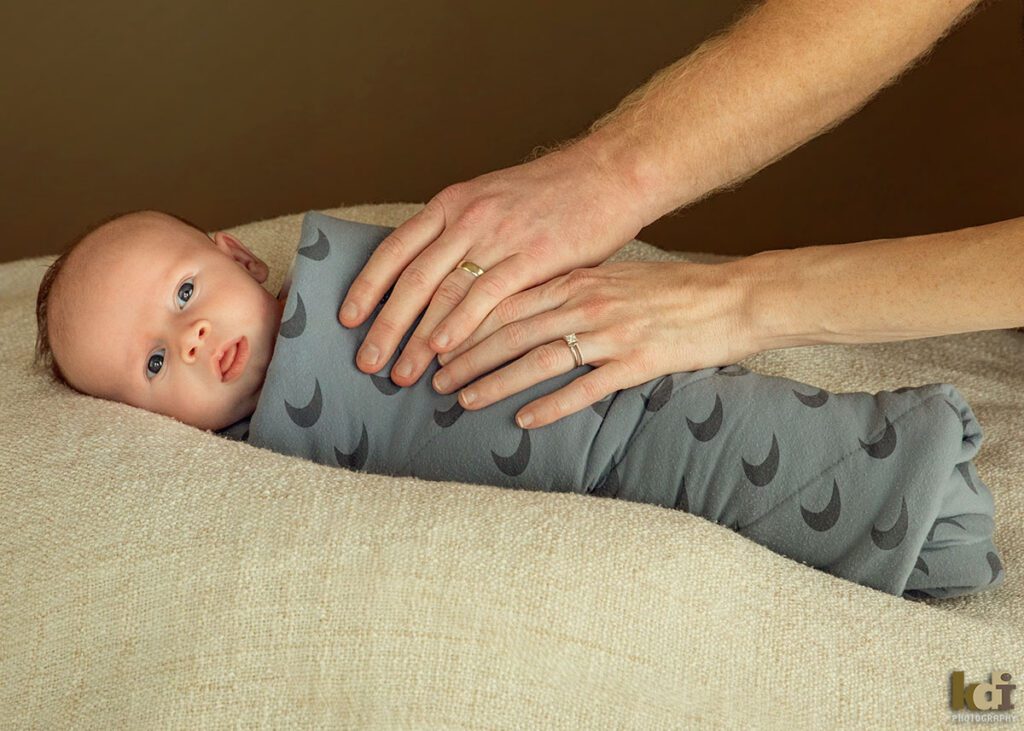 A mother and father's hands are seen holding a newborn baby boy who is wrapped in blue swaddle blanket, burrito style, color newborn portrait of a new family in a photo studio in Flagstaff by newborn and family photographer, KDI Photography