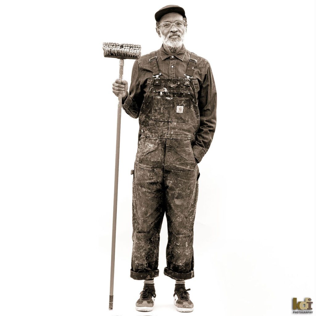 Black and white portrait of artist Chip Thomas, portrayed against a white background, holding his wheat-paste brush, wearing glasses, a hat and his painting overalls, portrait by Flagstaff Photographer, KDI Photography.